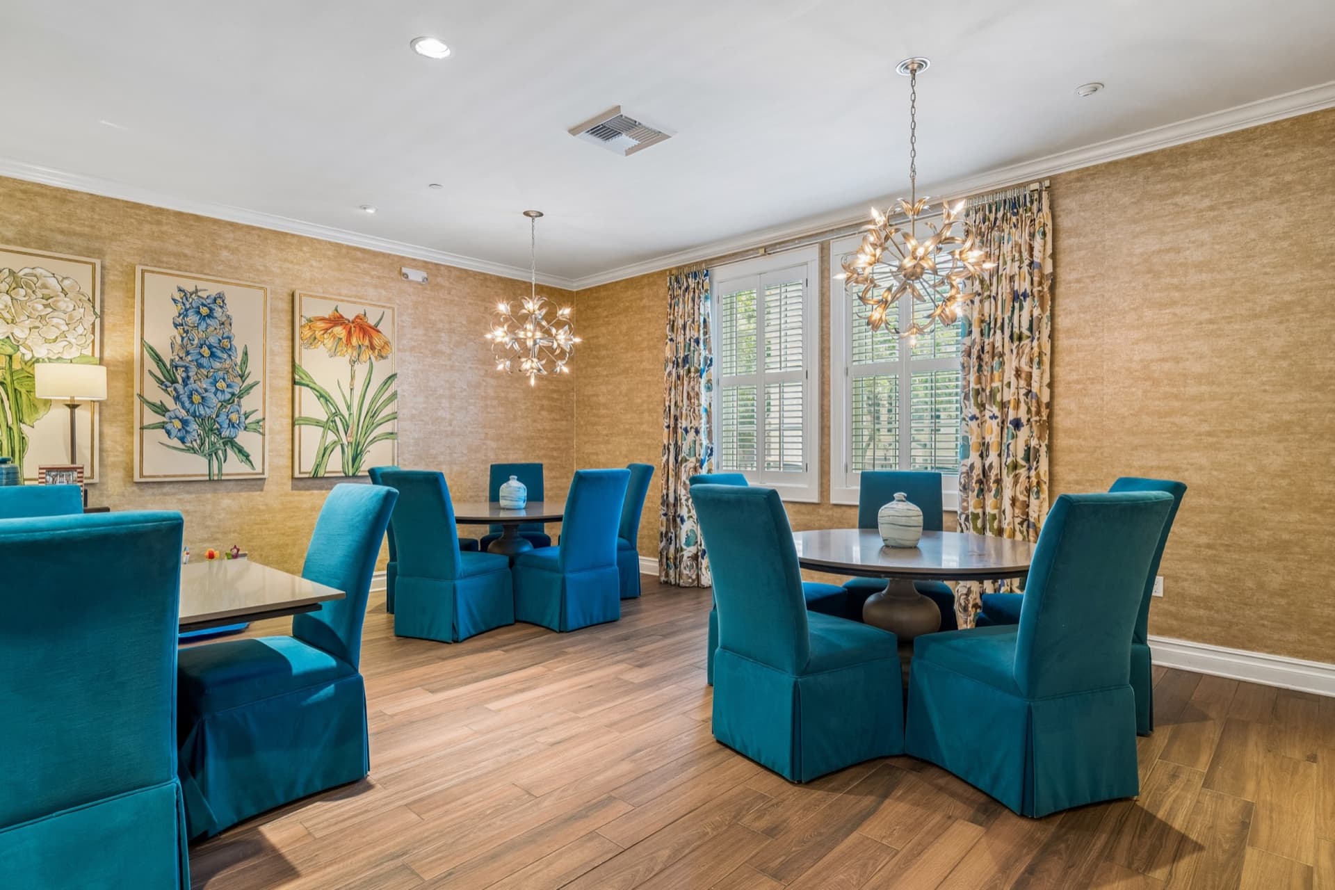 Community dining room with round tables, teal upholstered chairs, chandeliers, and botanical artwork.