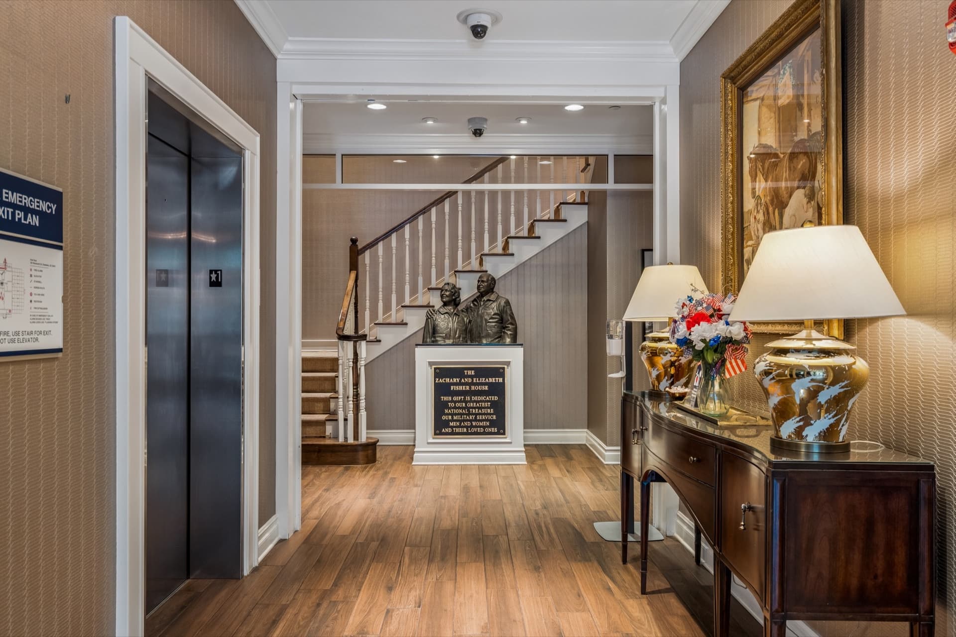 Foyer of the Charleston Fisher House with a staircase, elevator, and a bronze statue of Zachary and Elizabeth Fisher above a dedication plaque.