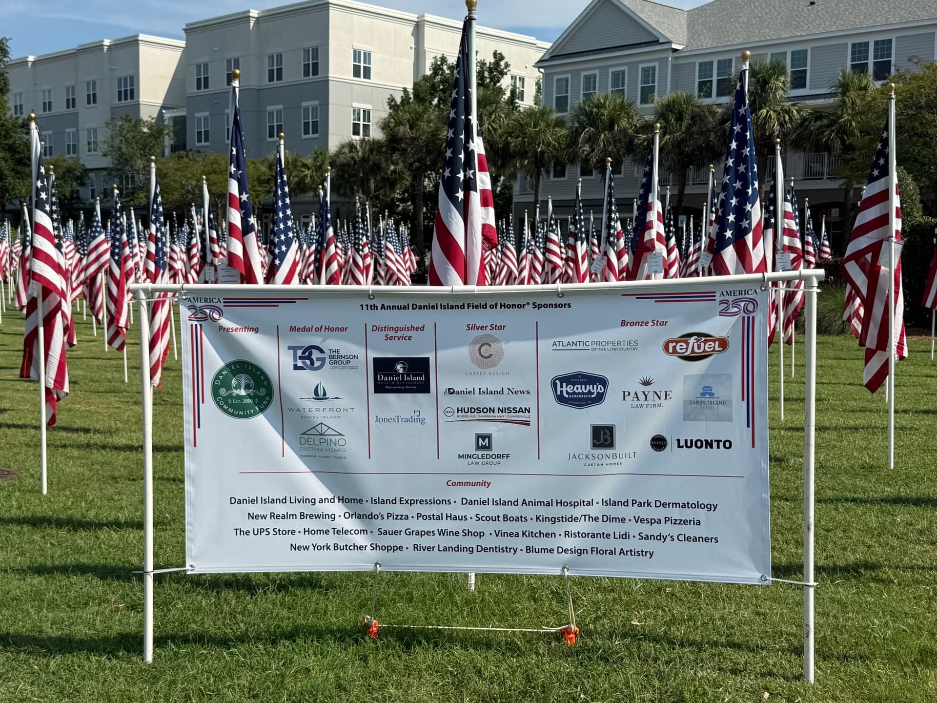 A white-framed banner listing the 11th Annual Daniel Island Field of Honor sponsors, with rows of small American flags planted in the grass behind it