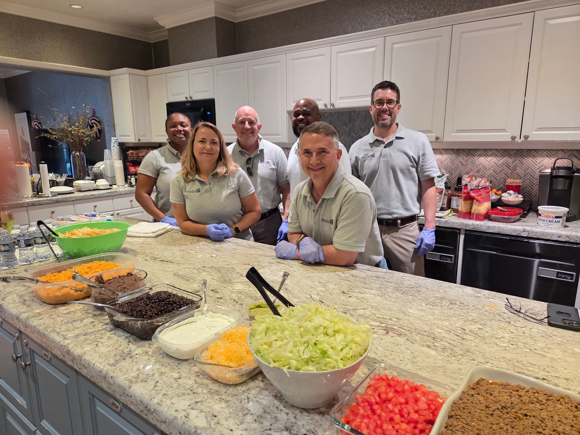 Five Modus21 volunteers standing behind the Fisher House kitchen island, with taco bar fixings of lettuce, cheese, beans, tomatoes, and seasoned beef laid out on the counter in front of them
