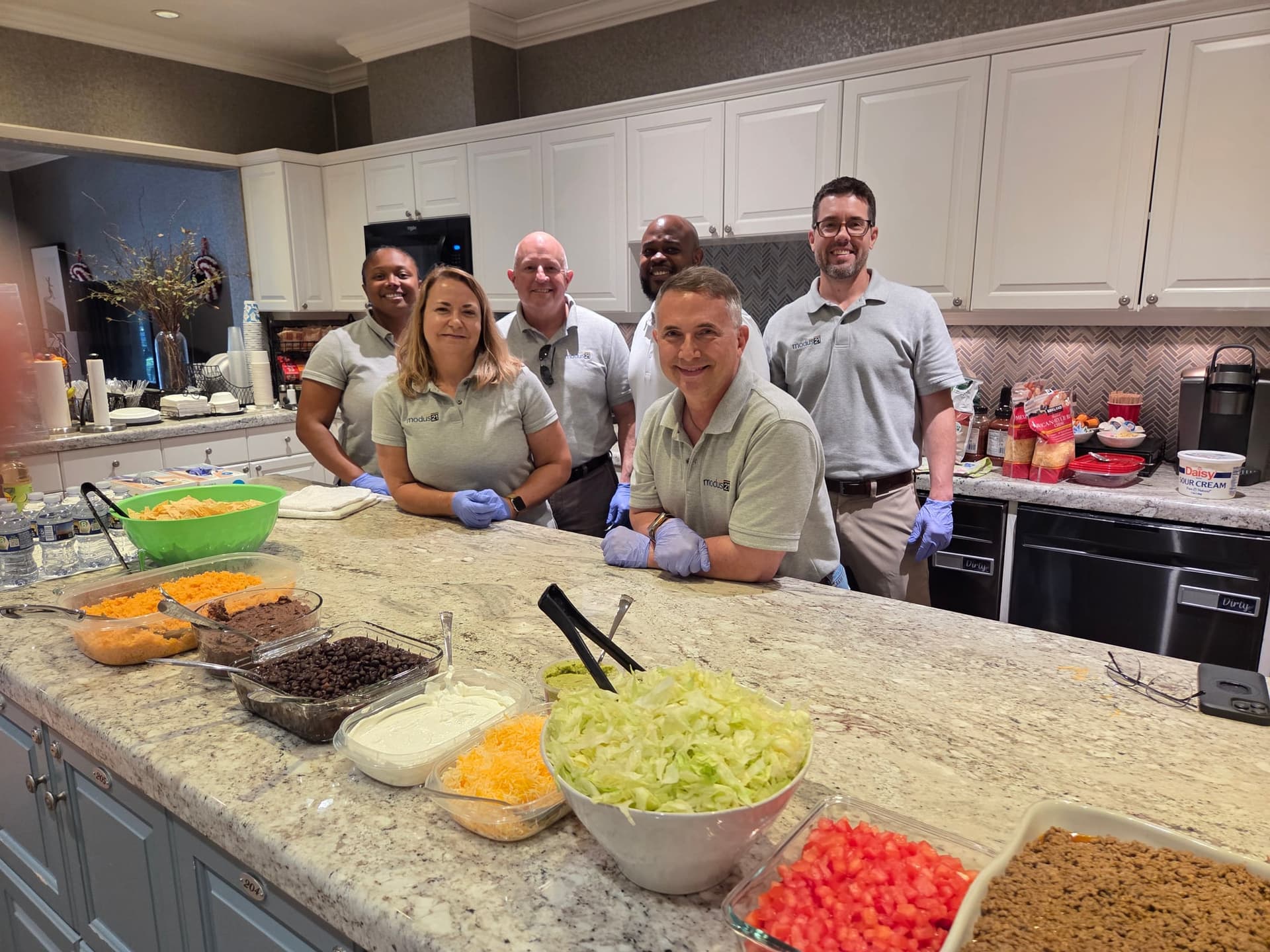 Five Modus21 volunteers standing behind the Fisher House kitchen island, with taco bar fixings of lettuce, cheese, beans, tomatoes, and seasoned beef laid out on the counter in front of them