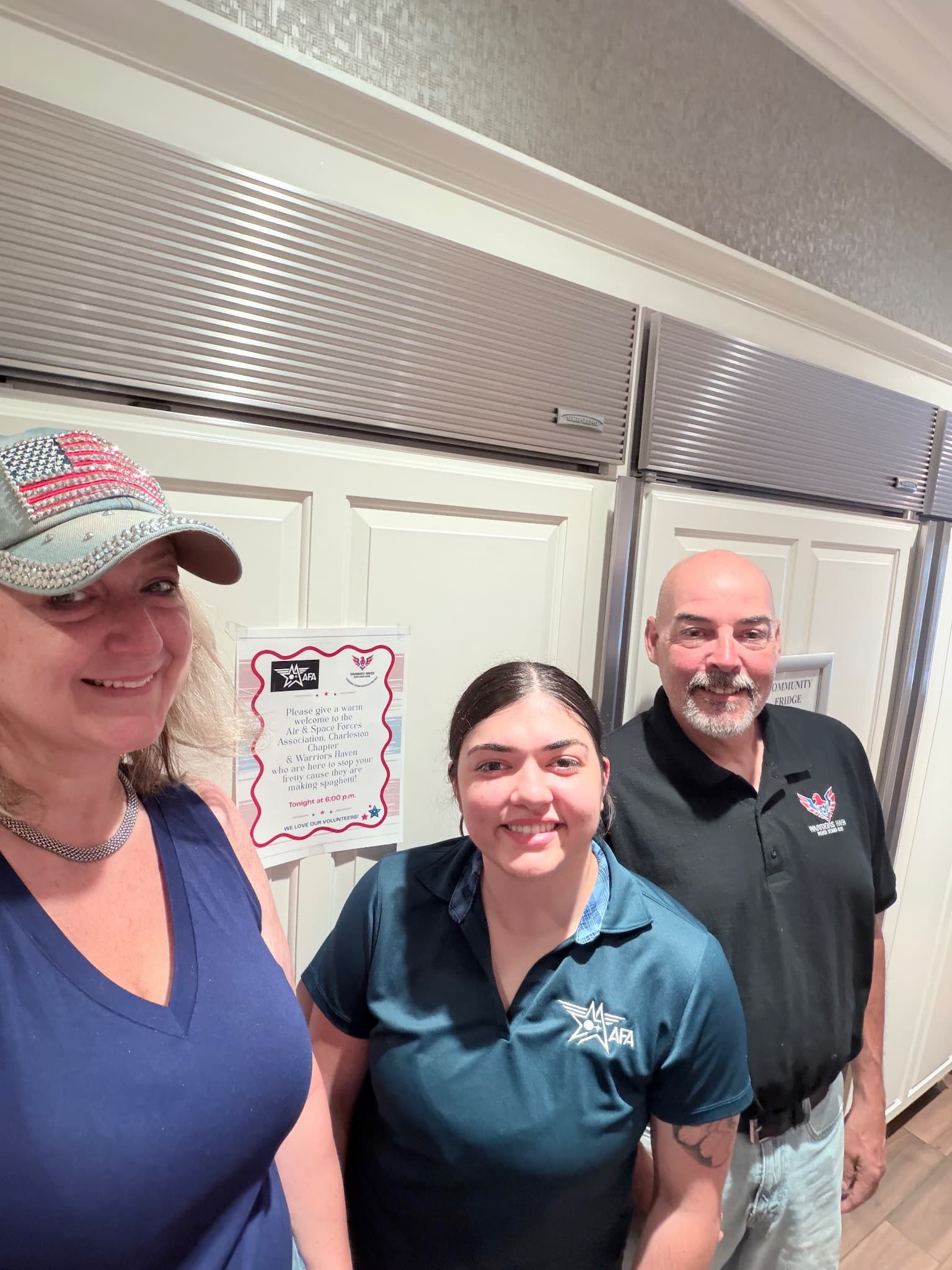 Three volunteers from Air & Space Forces Association Charleston and Warriors Haven smiling in the Fisher House beside a printed sign welcoming them for spaghetti night