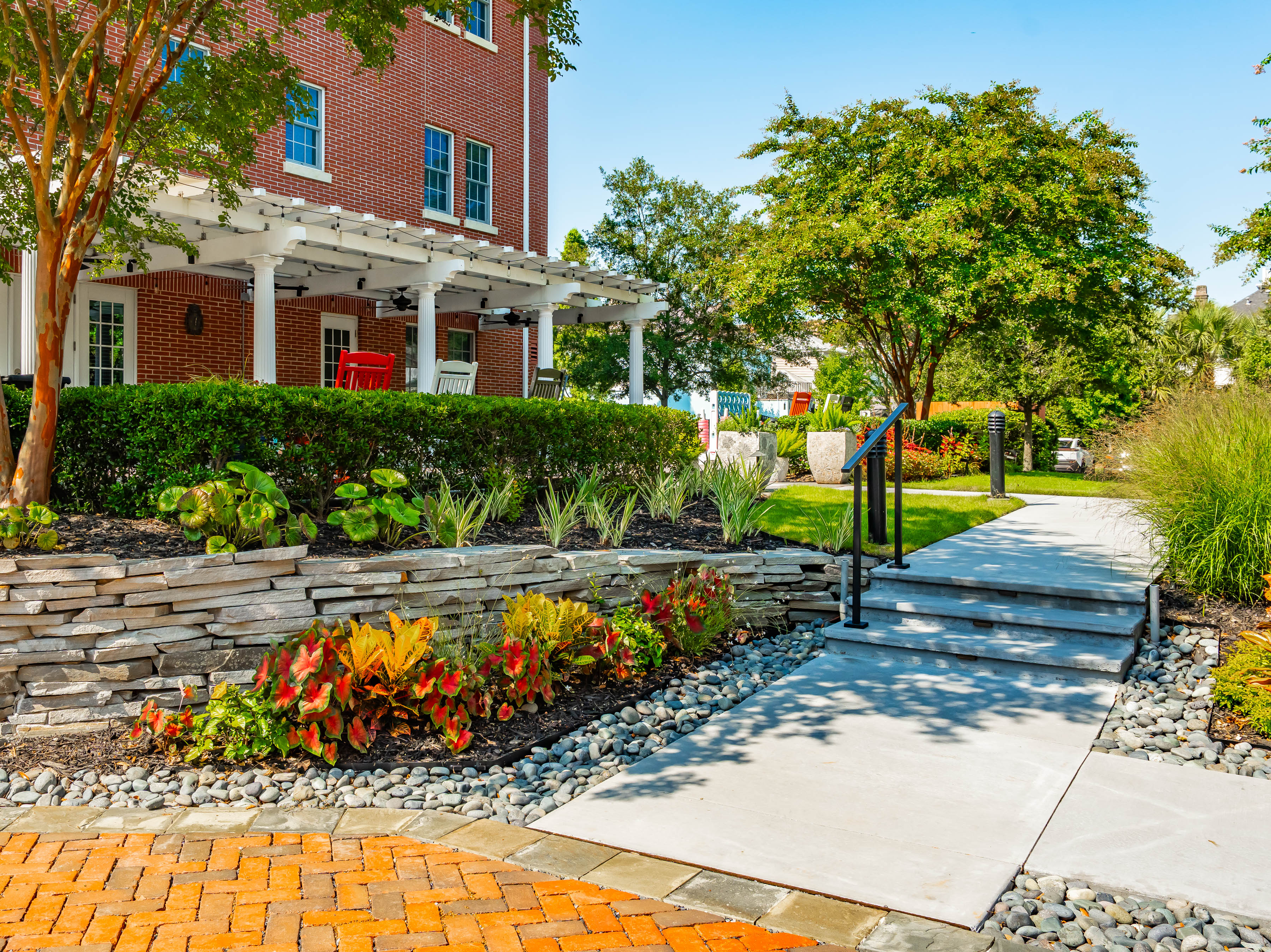 The landscaped front walkway of the Charleston Fisher House, with a stacked-stone planting bed, colorful caladiums, and rocking chairs under the white pergola