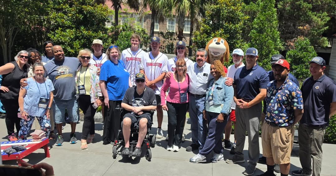 Charleston RiverDogs players in white jerseys, the Charlie T. RiverDog mascot, veteran families, and volunteers gathered together outdoors at the Charleston Fisher House