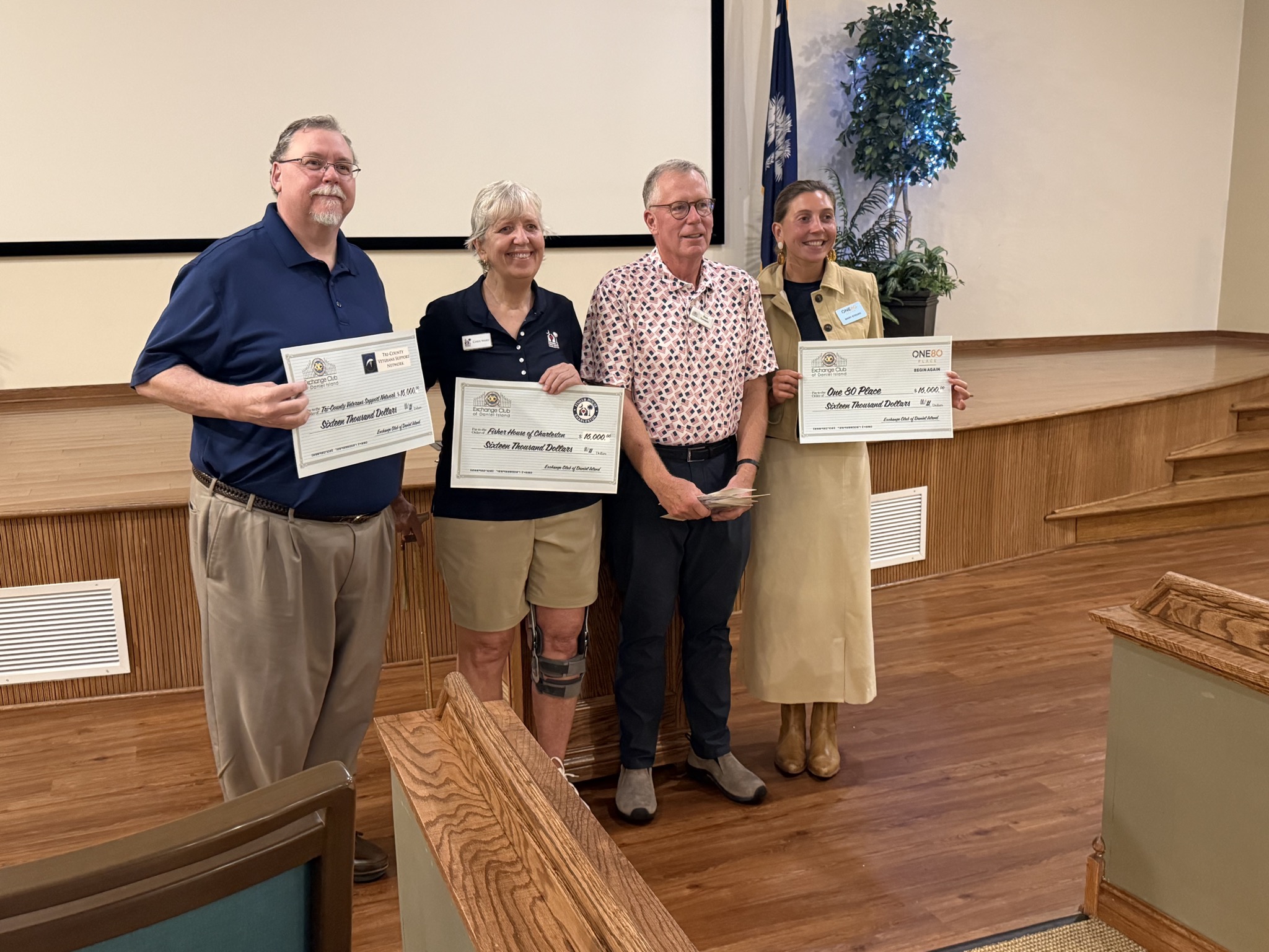Four people standing in a meeting hall holding oversized Exchange Club of Daniel Island checks for $16,000 each, made out to the Tri-County Veterans Support Network, Fisher House of Charleston, and One80 Place