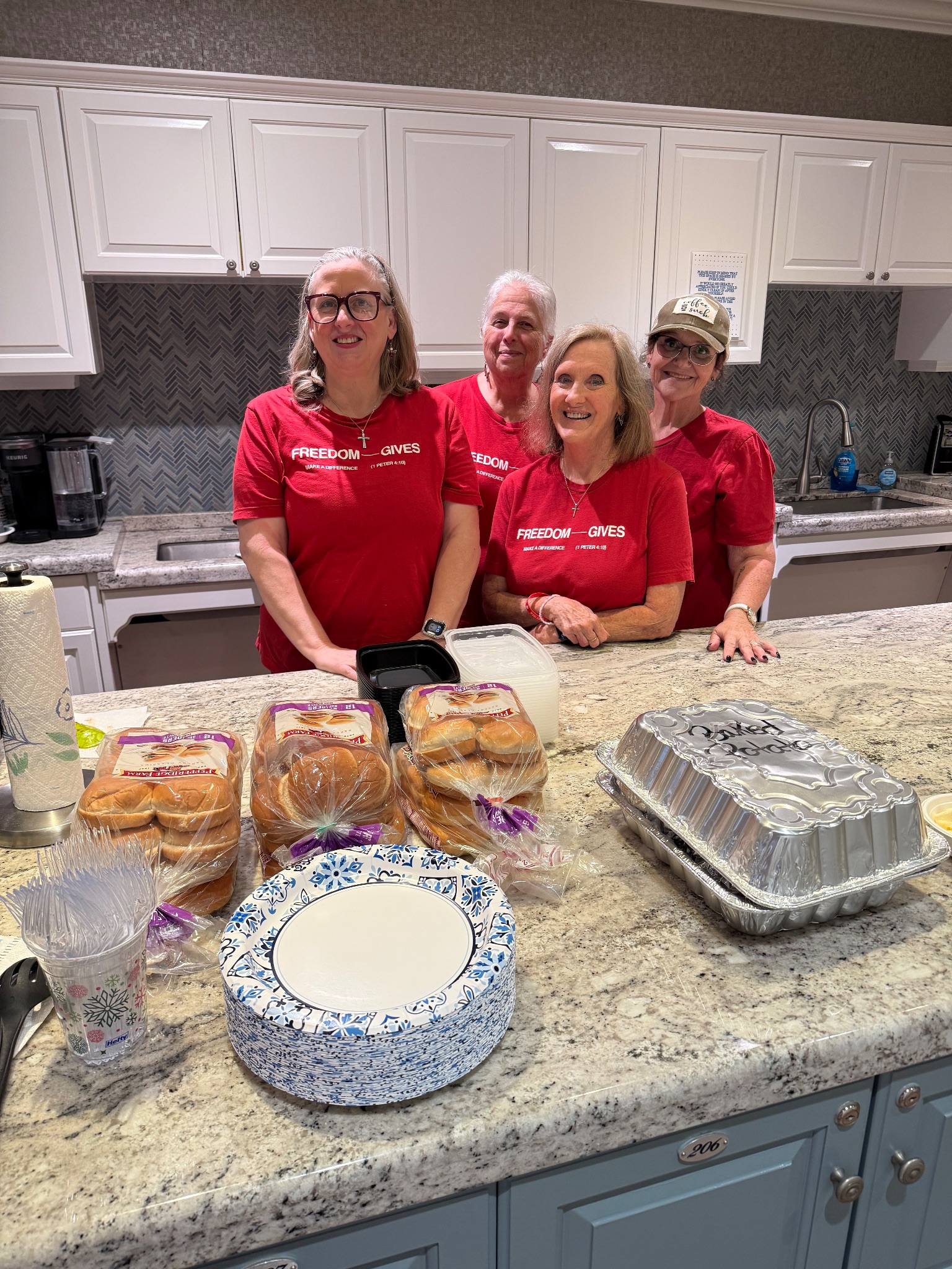 Four Freedom Church volunteers in red FREEDOM GIVES T-shirts smiling behind the Fisher House kitchen island, with packages of hamburger buns, a foil pan of baked potatoes, and a stack of paper plates laid out in front of them