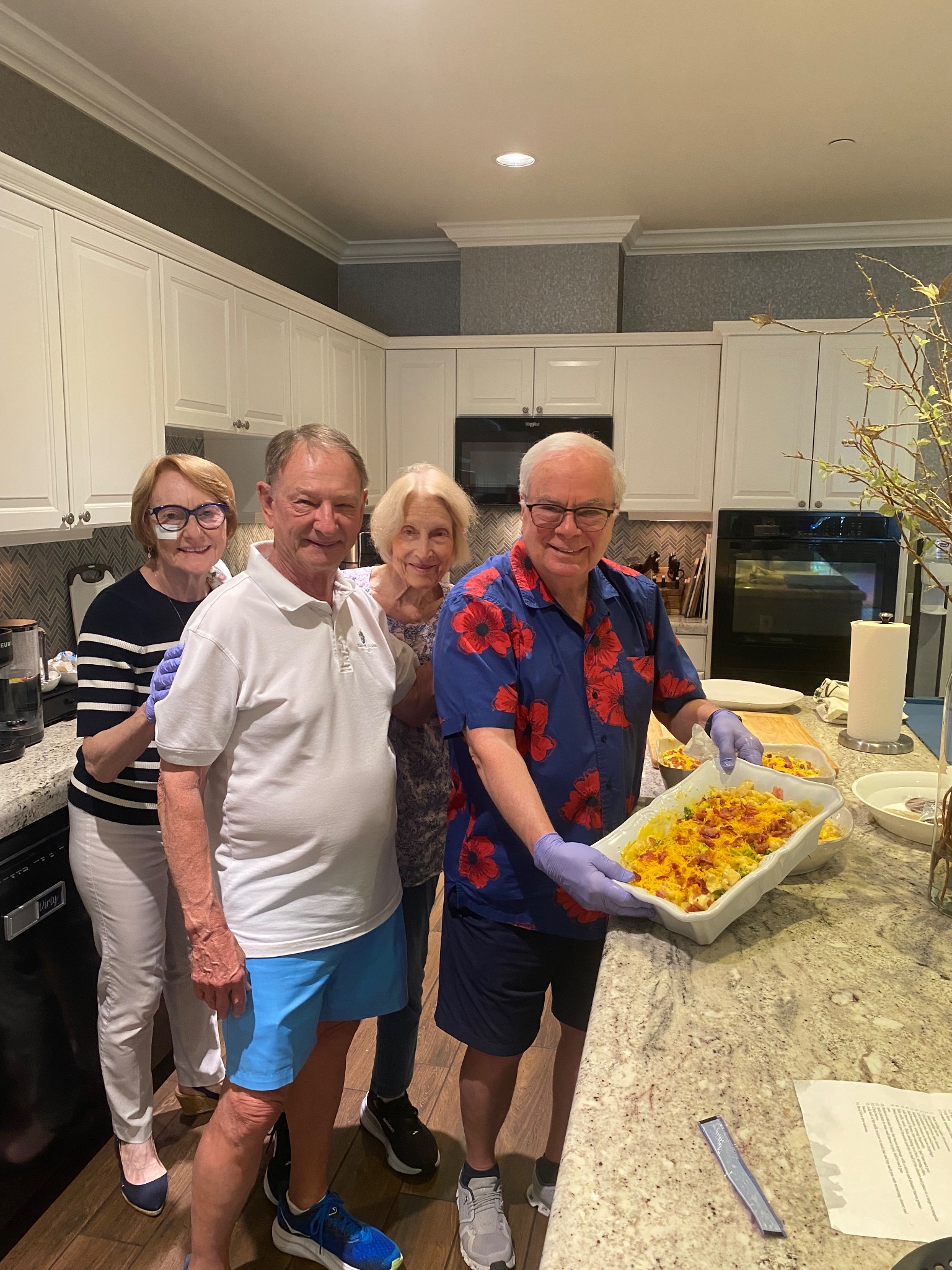 Four Grace Church Cathedral volunteers smiling in the Fisher House kitchen, one holding a freshly prepared casserole topped with cheese and bacon