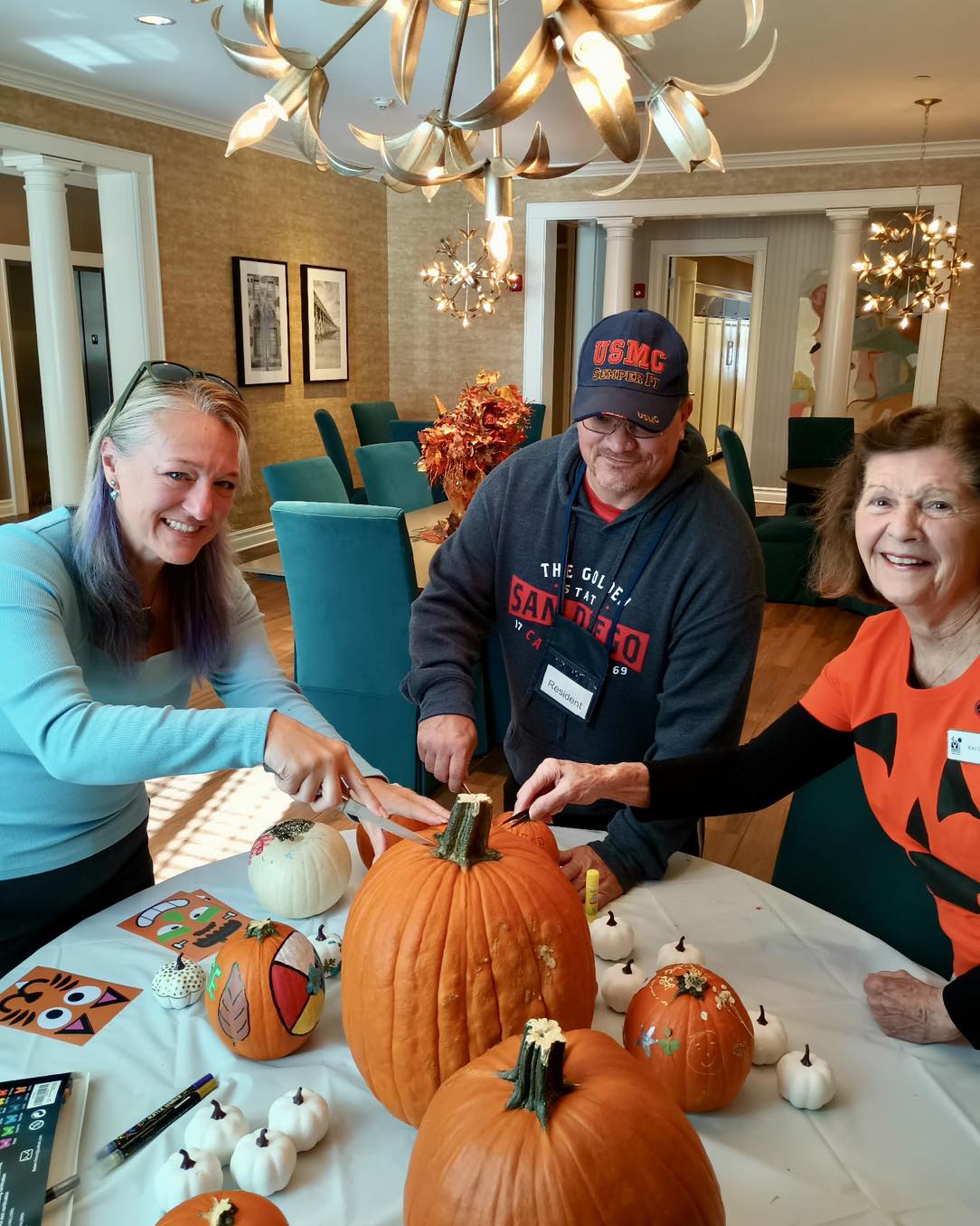Two volunteers and a Fisher House guest in a USMC cap carving and decorating pumpkins together at a table in the Fisher House common room