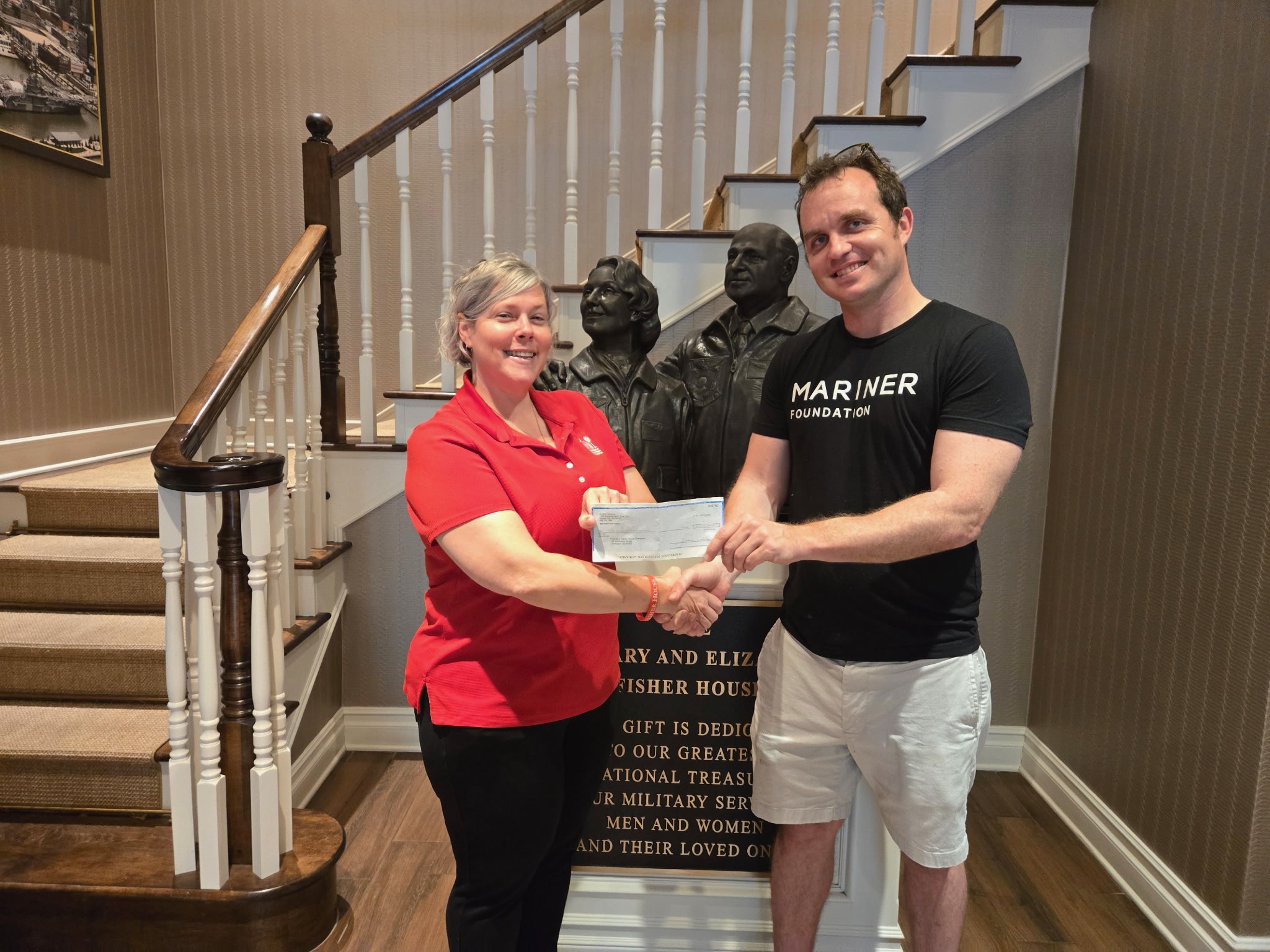 A Friends of Fisher House Charleston staff member in a red polo and a Mariner Foundation team member shaking hands while holding a $2,500 check in front of the Fisher House bronze sculpture and dedication plaque