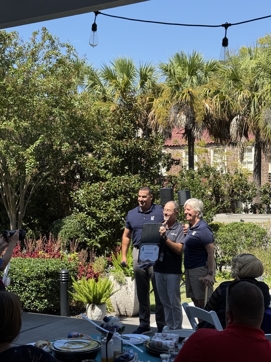 Jonathan Kessler holding an open certificate of appreciation with a blue ribbon on it, standing on the Fisher House patio between a man in a navy polo and Friends of Fisher House Charleston Vice President Chris Ward