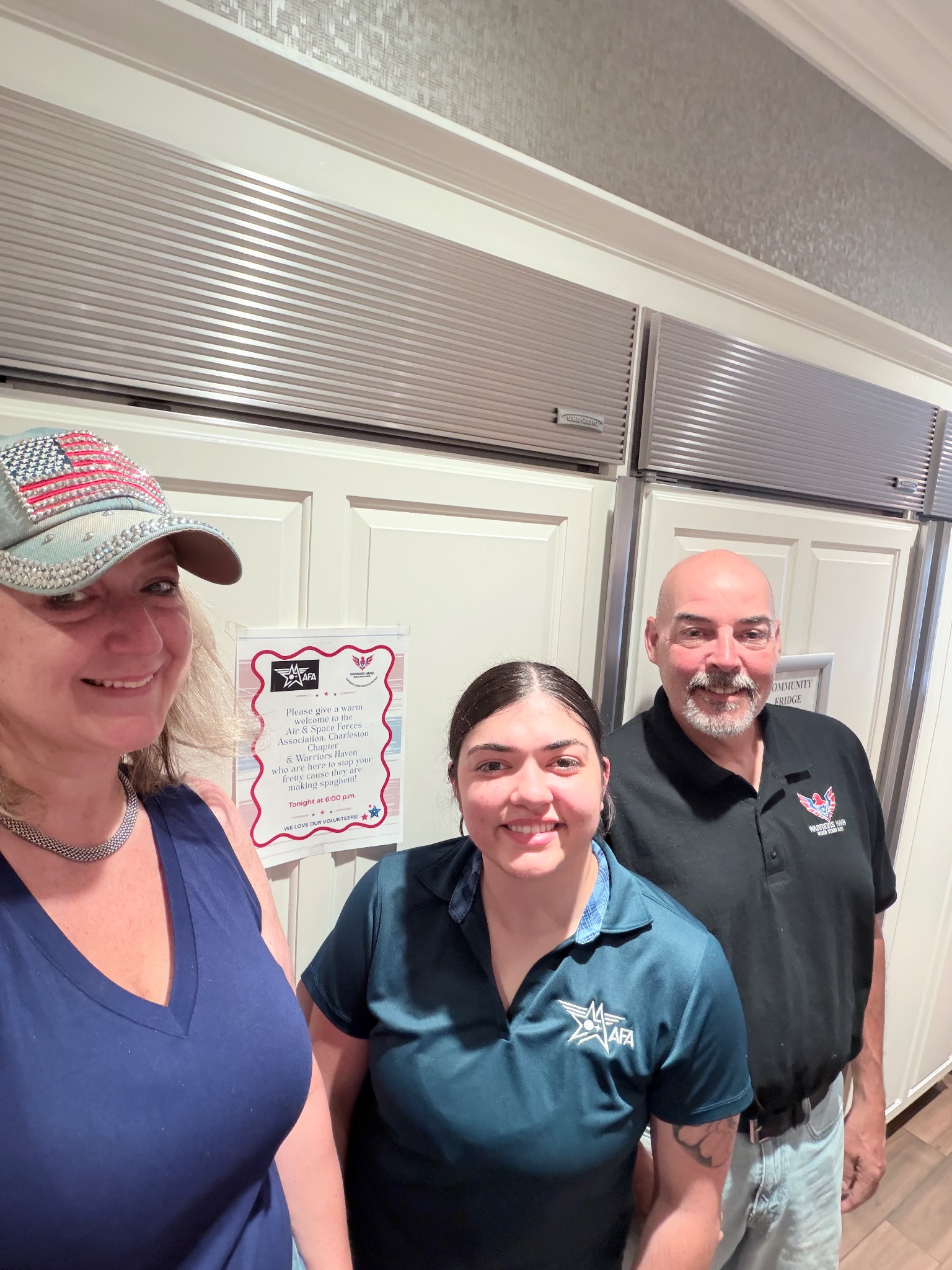 Three volunteers from Air & Space Forces Association Charleston and Warriors Haven smiling in the Fisher House beside a printed sign welcoming them for spaghetti night