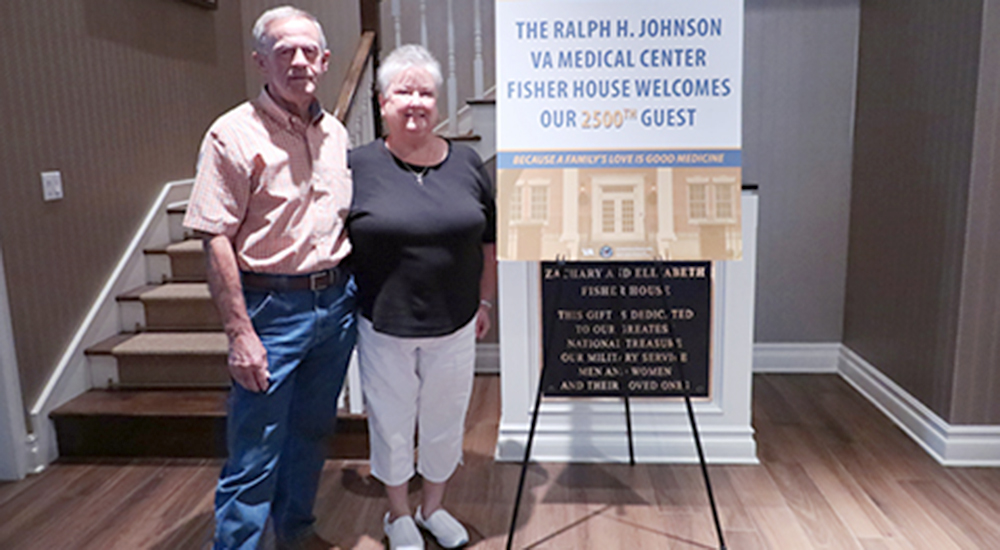 A veteran and his wife standing beside a poster welcoming the Charleston Fisher House's 2,500th guest family