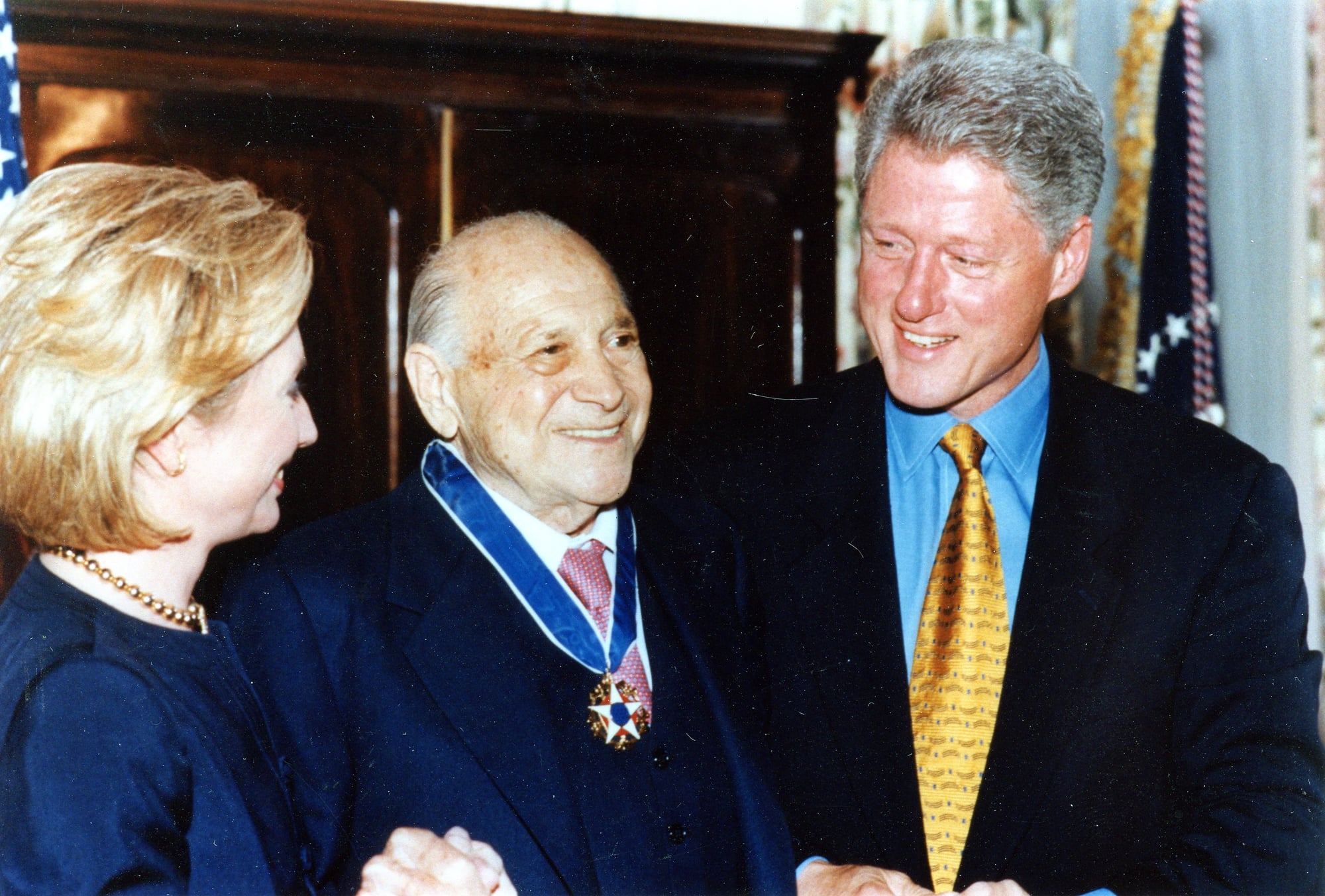 Zachary Fisher wearing the Presidential Medal of Freedom, smiling between President Bill Clinton and First Lady Hillary Clinton at the 1998 White House ceremony